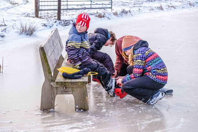 Een warm en vrolijk wintermoment op natuurijs: kinderen die samen schaatsen onderbinden, klaar voor nieuwe rondjes over het bevroren weiland. De kleurrijke mutsen en jassen, het houten bankje en de witte ijsvlakte vormen samen een typisch Hollands tafereel vol gezelligheid en winterplezier