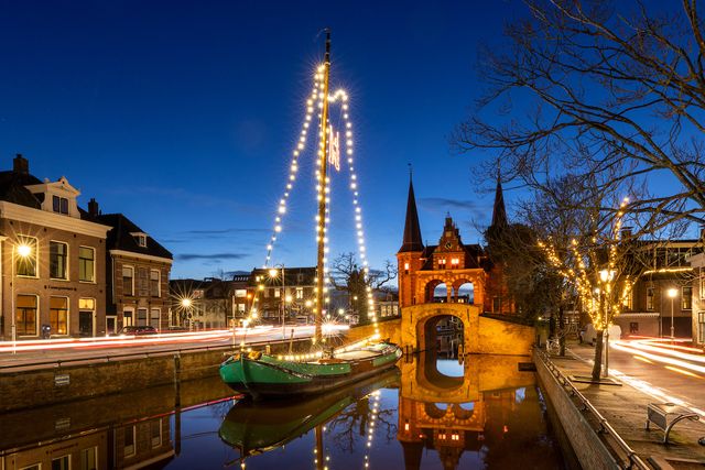 De Waterpoort in Sneek in het donker met op de voorgrond in het water een skûtsje met lampjes in de mast.