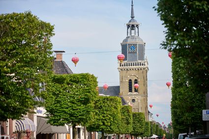 De kerktoren van de Midstraat in Joure. Onder te toren een lange rij met grote vierkante bomen en in de lucht hangen Chinese lampionnen.
