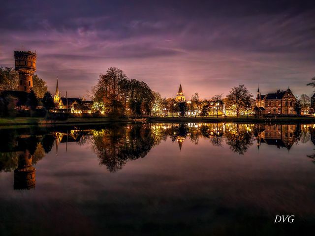 De binnenstad van Woerden bij avondlicht, met de watertoren, kerktorens en historische panden die sfeervol weerspiegelen in het stille water van de Singel.
