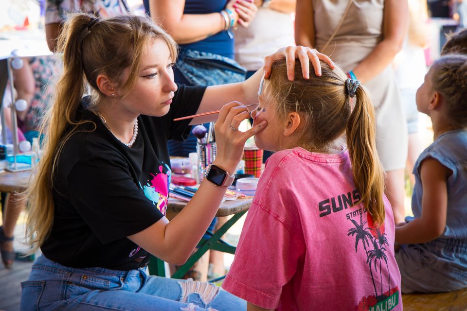 Kinderactiviteit met schminken tijdens Festival ’t Wonder in Woerden, Groene Hart. Schminkster beschildert het gezicht van een kind op een familiefestival met creatieve kinderactiviteiten, entertainment en zomerse festivalsfeer.