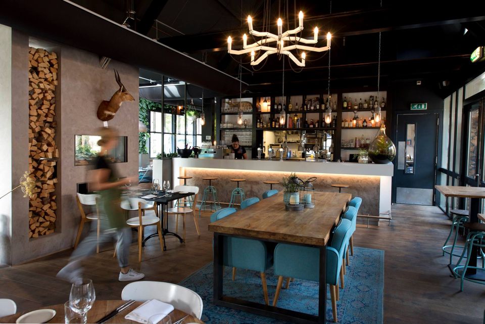 Restaurant Bistronoom Woonkamer in Woerden, Groene Hart, modern restaurantinterieur met bar, houten tafels, blauwe stoelen, open haard en sfeervolle hanglampen tijdens diner in eigentijdse horecagelegenheid.