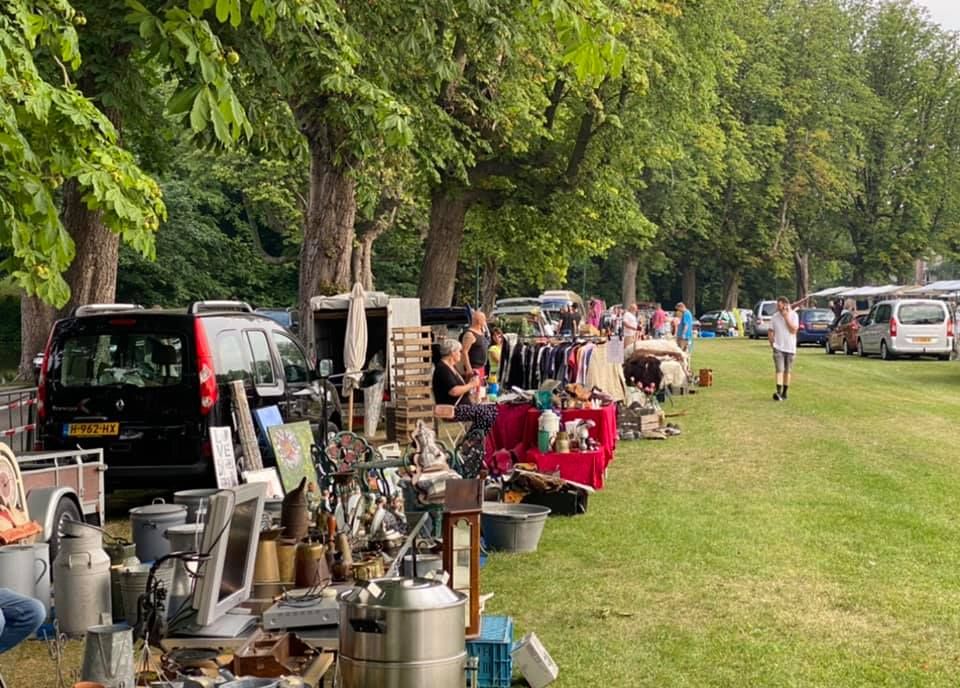 Vlooienmarkt in Woerden, Groene Hart, gezellige markt met kramen vol tweedehands spullen, antiek en kleding langs een met bomen omzoomd grasveld waar bezoekers tussen de koopjes struinen.