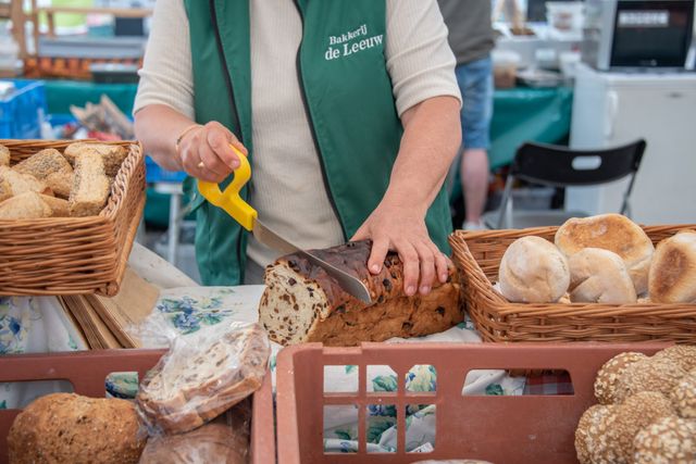 Markt met streekproducten Streekmarkt Woerden in Woerden, Groene Hart, kraam van Bakkerij de Leeuw waar een ambachtelijk rozijnenbrood wordt aangesneden tussen manden met versgebakken brood en harde broodjes.