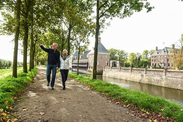 Een ouder stel wandelt langs de gracht bij Kasteel Amerongen, met het historische kasteel en de tuinen op de achtergrond. De foto laat wandelen, erfgoed en de groene omgeving van Amerongen op de Utrechtse Heuvelrug zien.