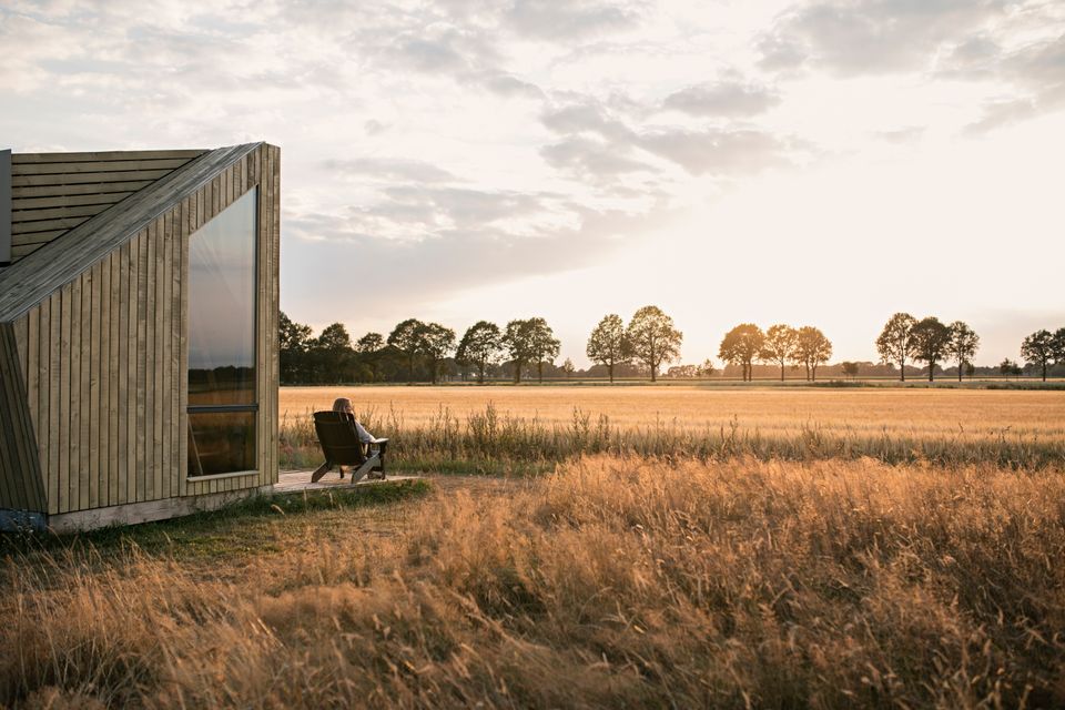 Een vrouw zit op een stoel voor een vakantiehuisje en kijkt uit over het korenveld.